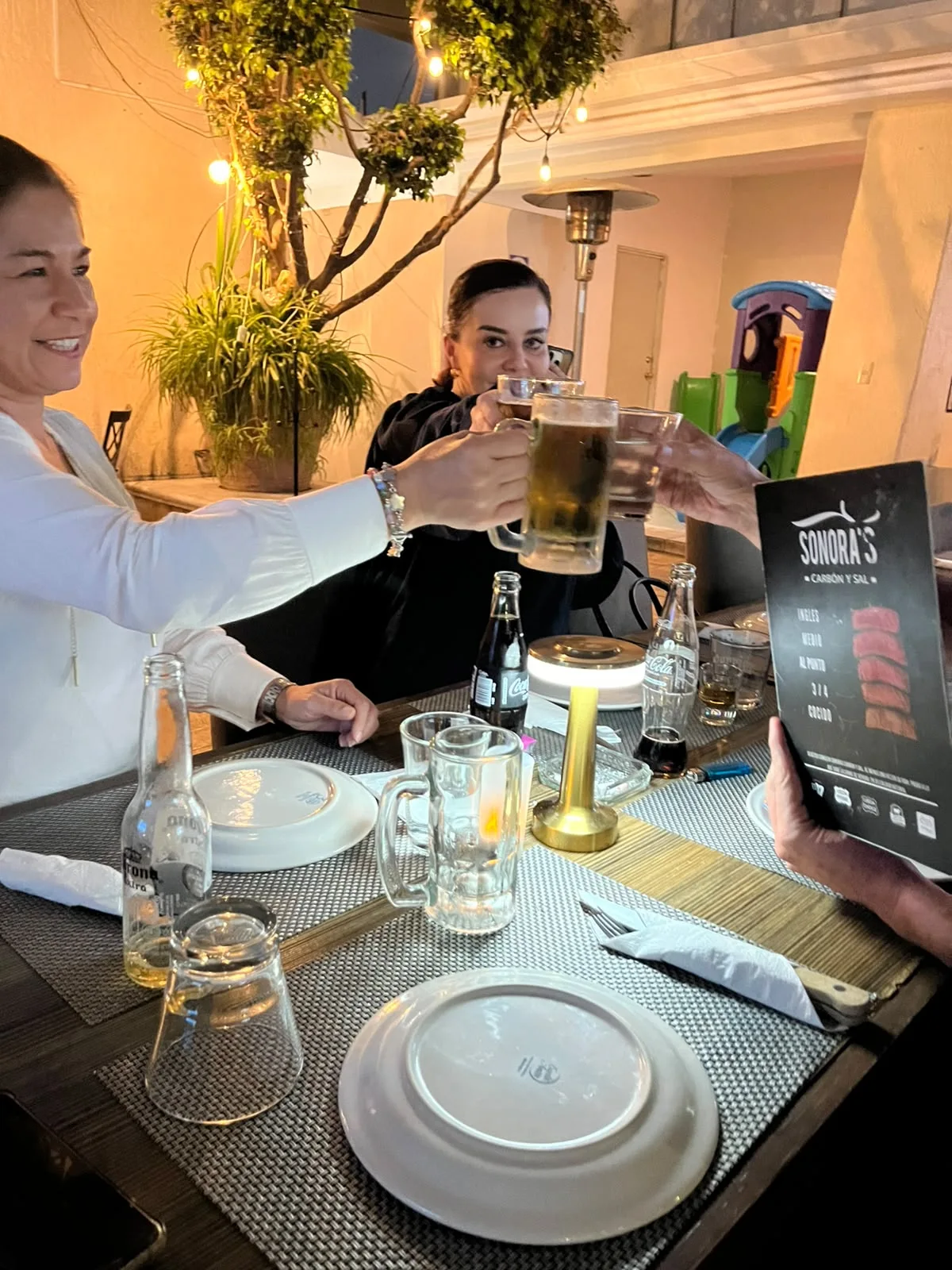 Brindis con cerveza y bebidas en mesa de Sonora's Carbón y Sal con el menú de cortes al carbón